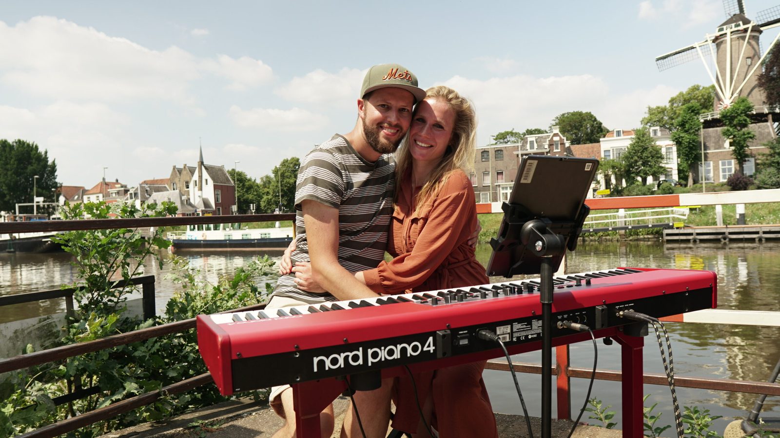 Zeb en Noesjka lachend achter een rode piano aan het water in Gouda, met een molen op de achtergrond